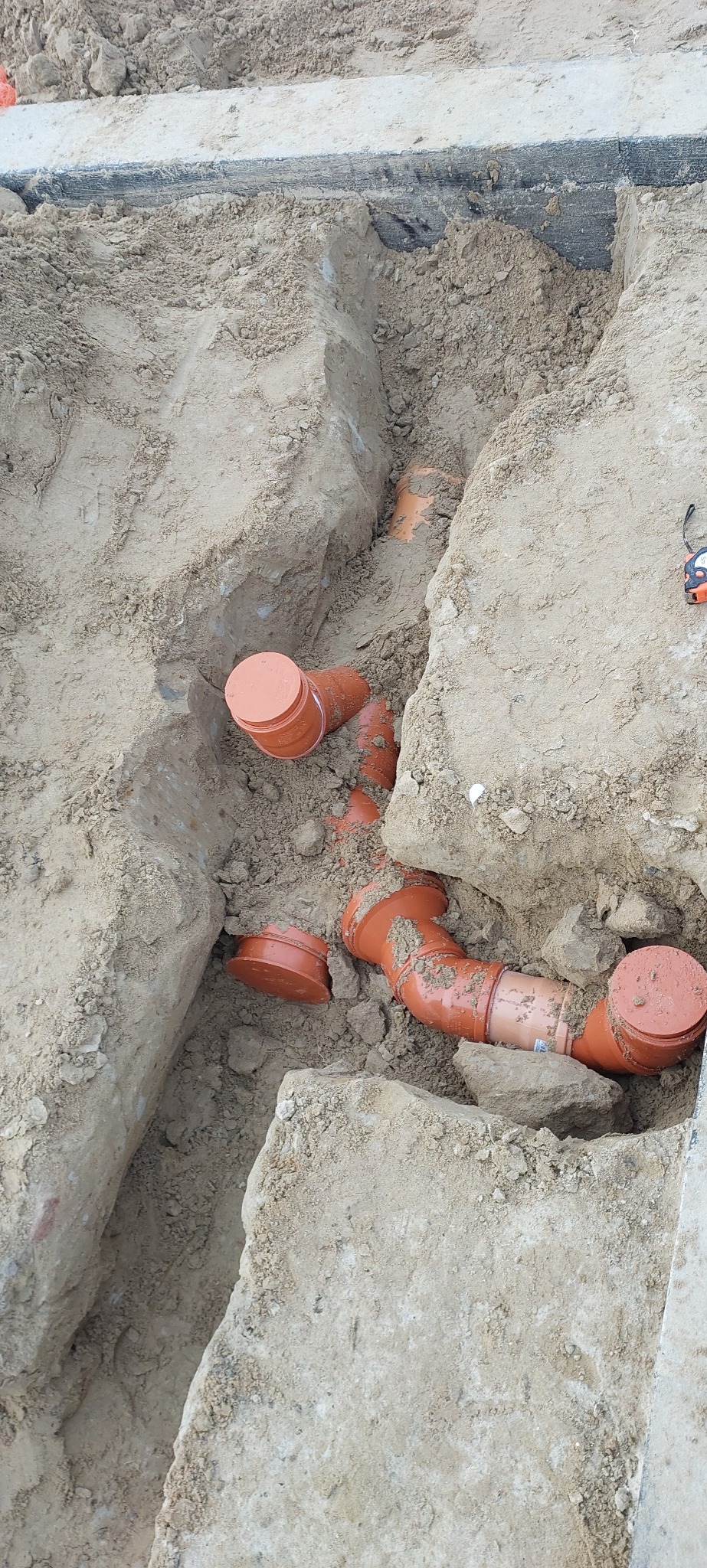 Pomarańczowe rury kanalizacyjne ułożone w wykopie ziemnym, widoczne z góry. Elementy instalacji połączone, przygotowane do zasypania.