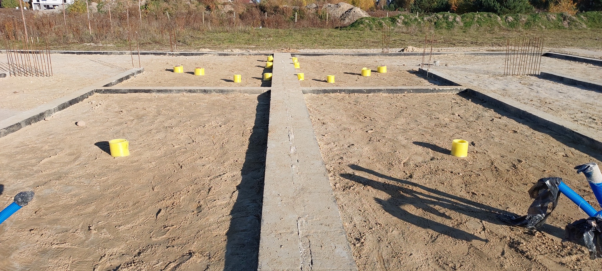 Fundamenty domu jednorodzinnego z żółtymi znacznikami, widoczne zbrojenia i częściowo zakopane rury. Ujęcie z góry, w słoneczny dzień.