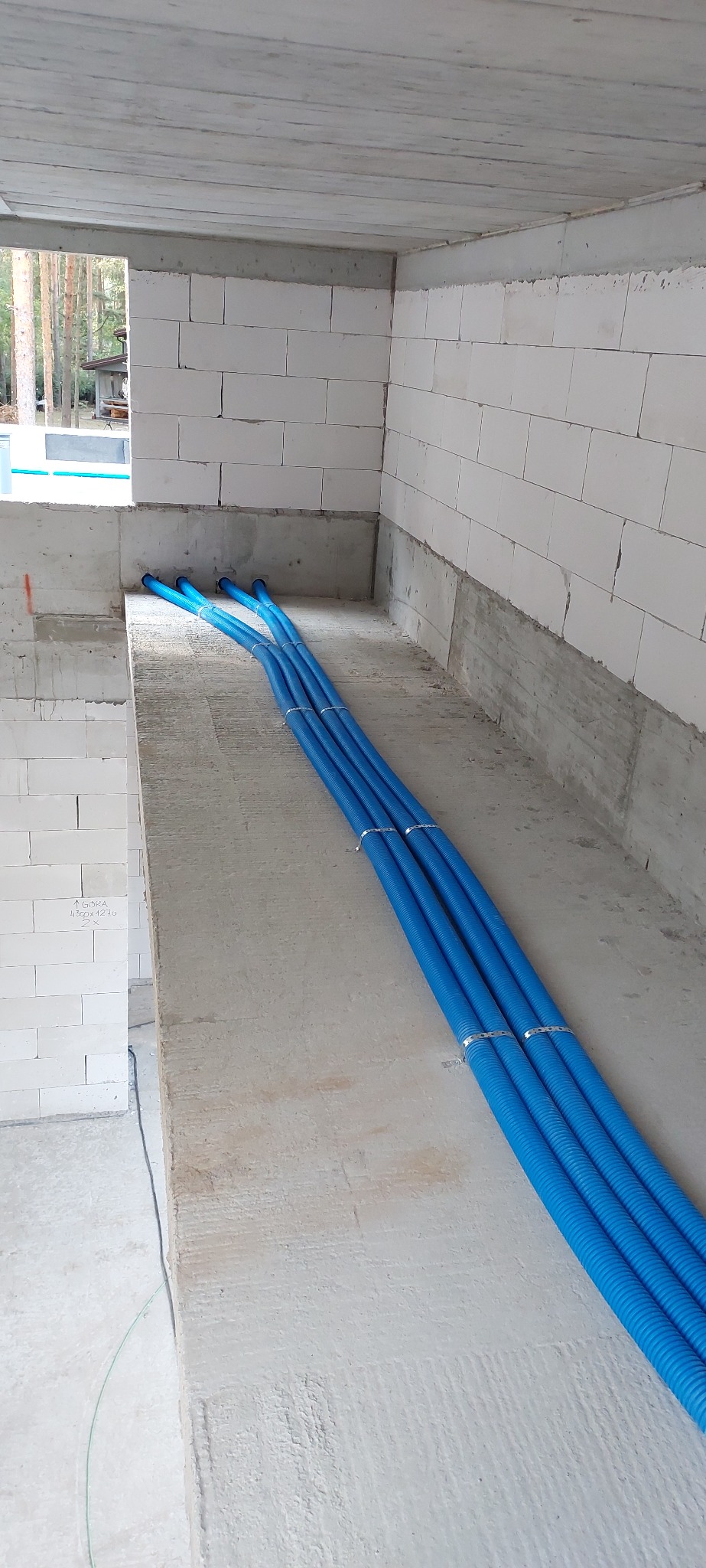 Niebieskie rury instalacyjne, przymocowane metalowymi obejmami, ułożone w surowym wnętrzu budynku z białych bloczków. Widoczne otwory w ścianie.