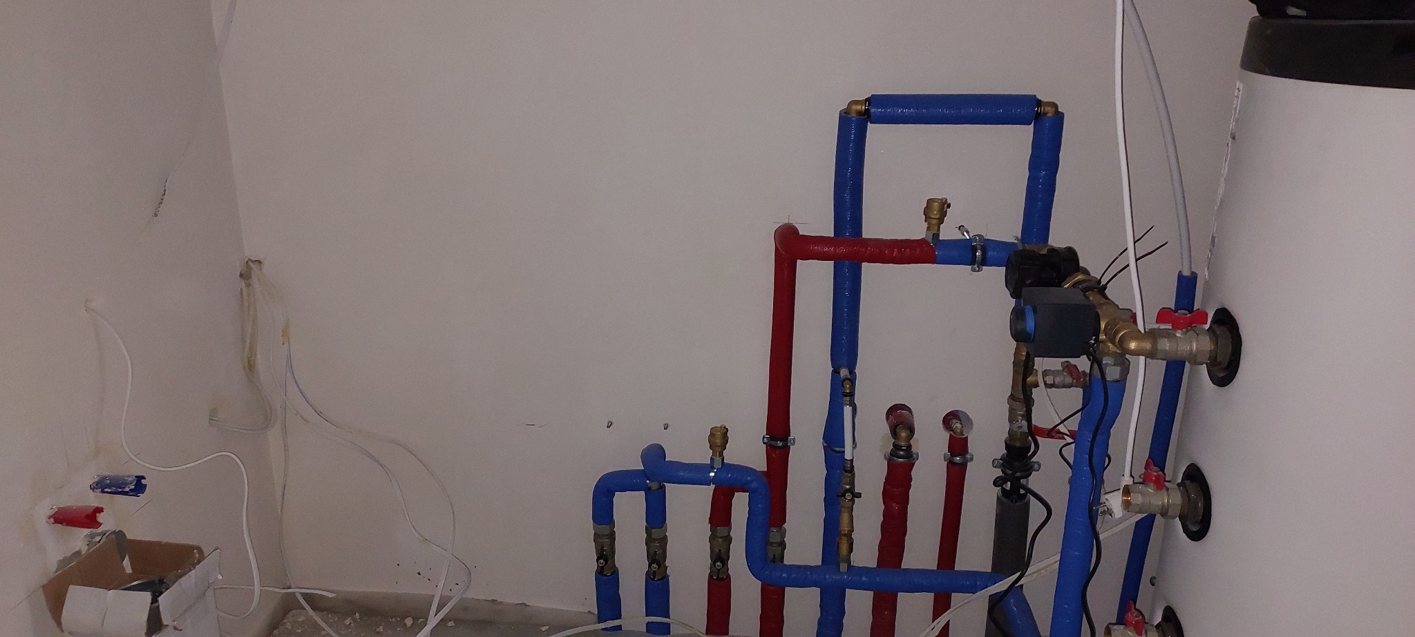 Instalacja grzewcza: rury w izolacji (czerwone i niebieskie) prowadzone wzdłuż ściany, z zaworami i pompą obiegową, obok biały zasobnik ciepłej wody.
