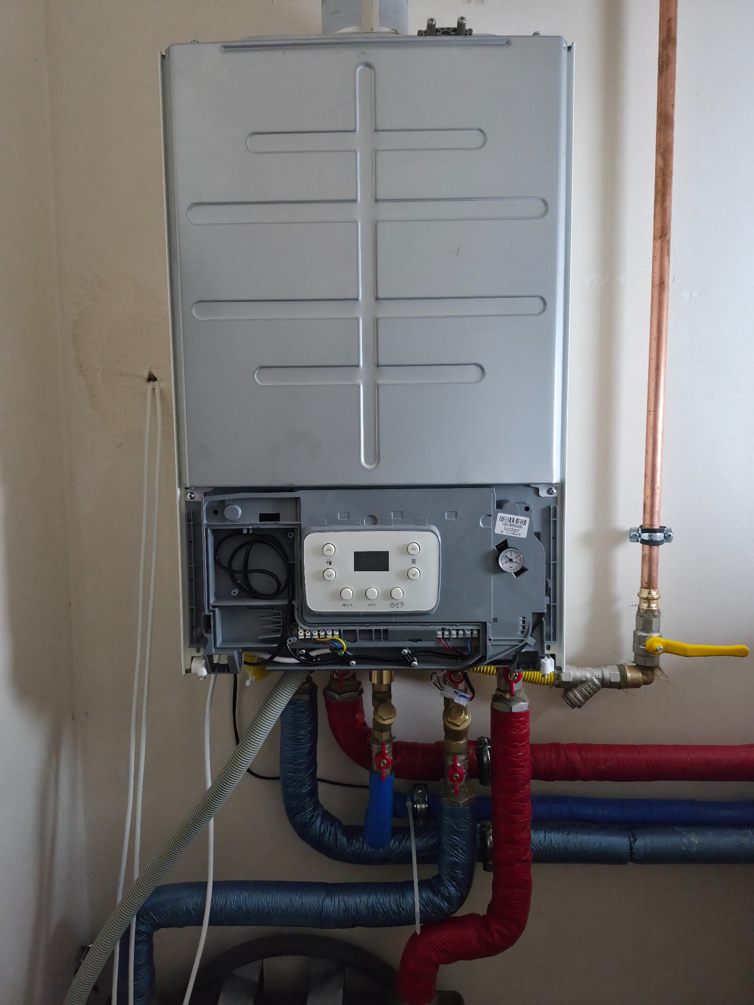 Nowoczesny piec gazowy dwufunkcyjny, z widocznymi rurami instalacji CO i CWU, zaworem gazowym z żółtą rączką i fragmentem ściany.