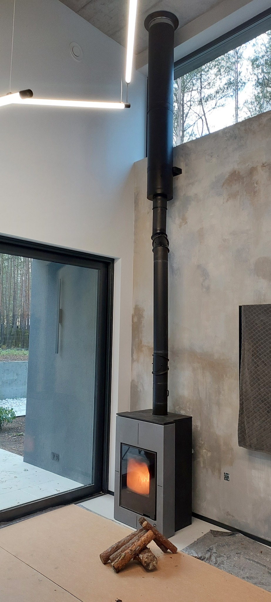 Nowoczesny piec wolnostojący z czarnym kominem w minimalistycznym wnętrzu. Widoczne płomienie i drewno obok pieca. Surowy beton na ścianie dodaje industrialnego charakteru.