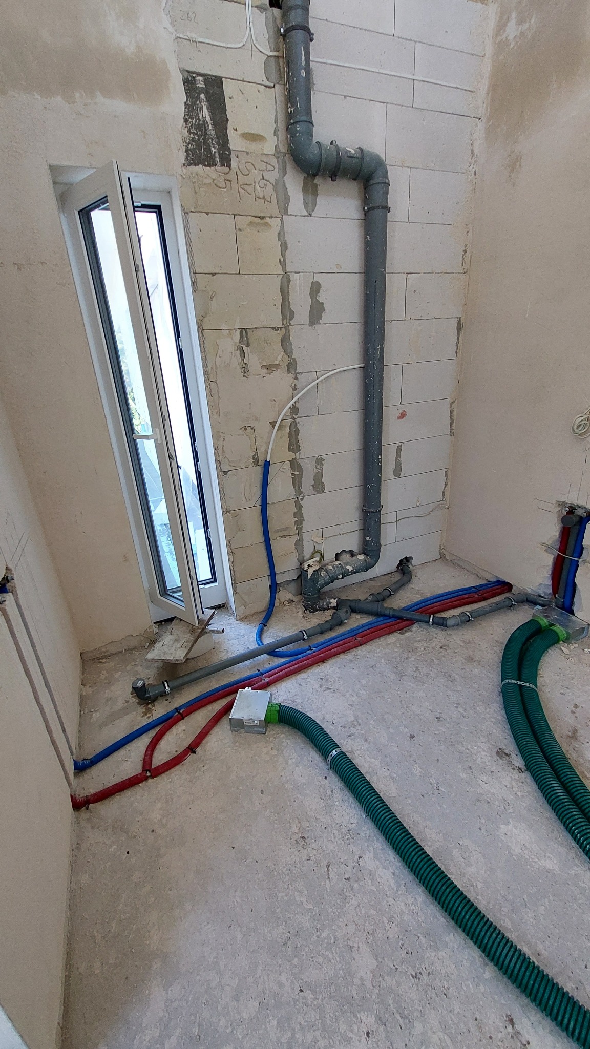 Surowy stan: rozprowadzone rury wodno-kanalizacyjne w kolorach niebieskim, czerwonym i zielonym na tle ściany z betonu komórkowego i otwartego okna.