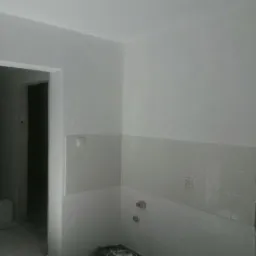 Wnętrze w trakcie remontu: ściany pomalowane na biało, częściowo wyłożone jasnymi płytkami, widoczne niedokończone wykończenia przy futrynie drzwi i surowy fragment ściany z odsłoniętym betonem.