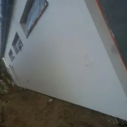 Boczny widok budynku z nową, białą elewacją. Okna zabezpieczone folią, teren wokół budynku przygotowany pod dalsze prace.