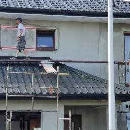 Pracownik na rusztowaniu podczas ocieplania elewacji domu jednorodzinnego, widoczne fragmenty izolacji z folią ochronną wokół okien, przygotowane płyty styropianowe oraz puszki z farbą na rusztowaniu.