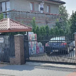 Dwóch pracowników na rusztowaniu przy domu w trakcie prac elewacyjnych, zabezpieczone okna folią, widoczne puszki z farbą, dom w stanie surowym, ogrodzenie z bramą w tle.