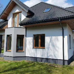 Świeżo otynkowana i pomalowana elewacja domu jednorodzinnego z ciemną dachówką, widoczny fragment trawnika i pracownik w kombinezonie przy wejściu.