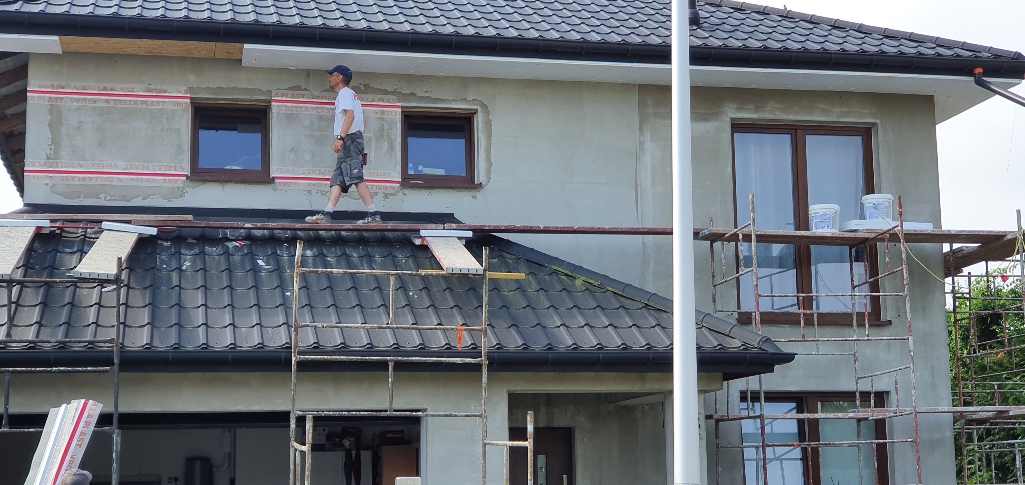 Pracownik na rusztowaniu podczas ocieplania elewacji domu jednorodzinnego, widoczne fragmenty izolacji z folią ochronną wokół okien, przygotowane płyty styropianowe oraz puszki z farbą na rusztowaniu.