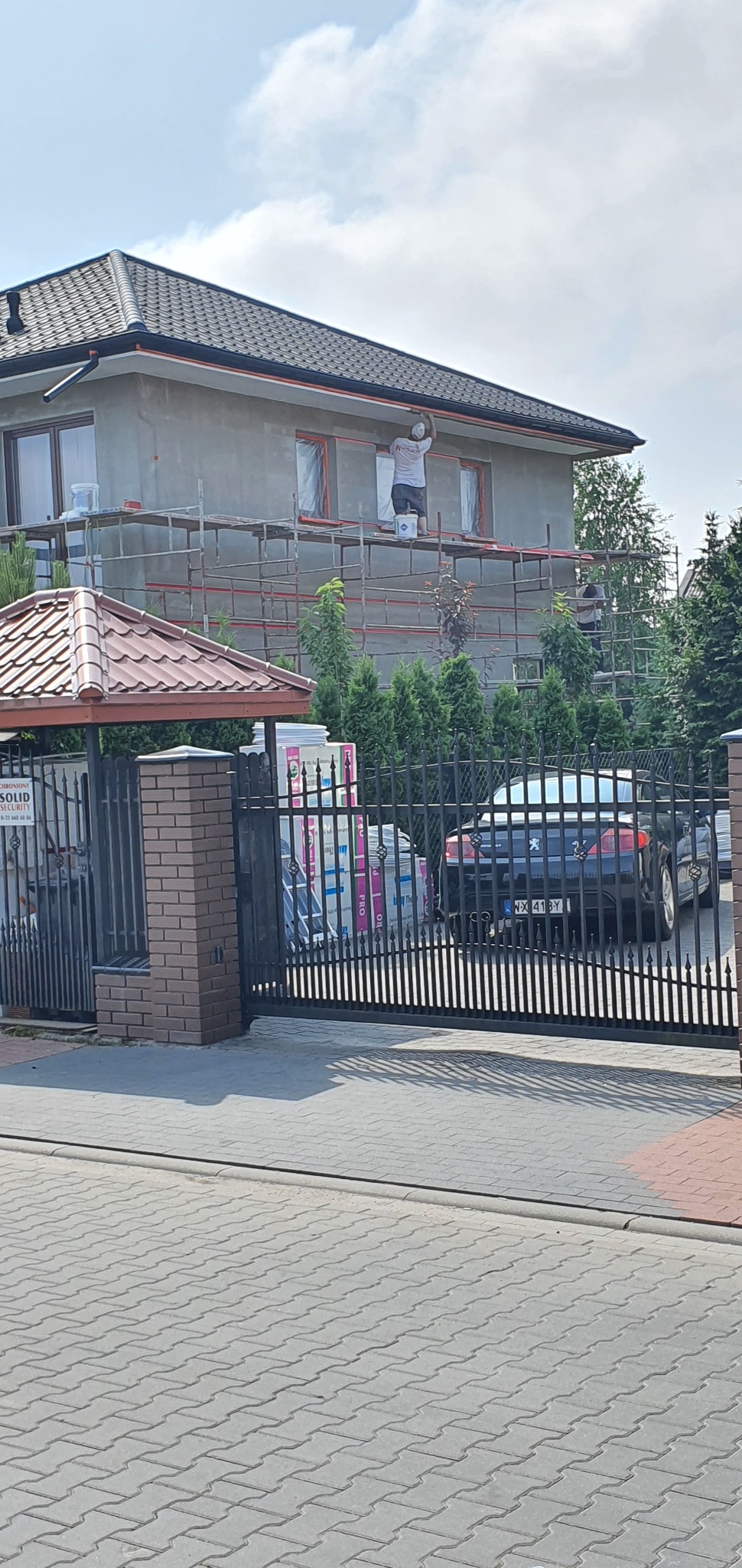 Dwóch pracowników na rusztowaniu przy domu w trakcie prac elewacyjnych, zabezpieczone okna folią, widoczne puszki z farbą, dom w stanie surowym, ogrodzenie z bramą w tle.
