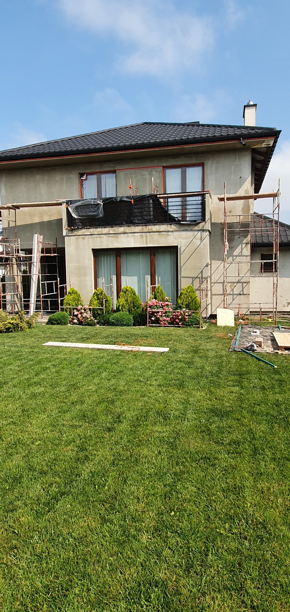 Dom w trakcie prac elewacyjnych, widoczne rusztowania, elewacja w szarym tynku, zabezpieczony balkon, zadbany trawnik z ozdobnymi krzewami i kwiatami.