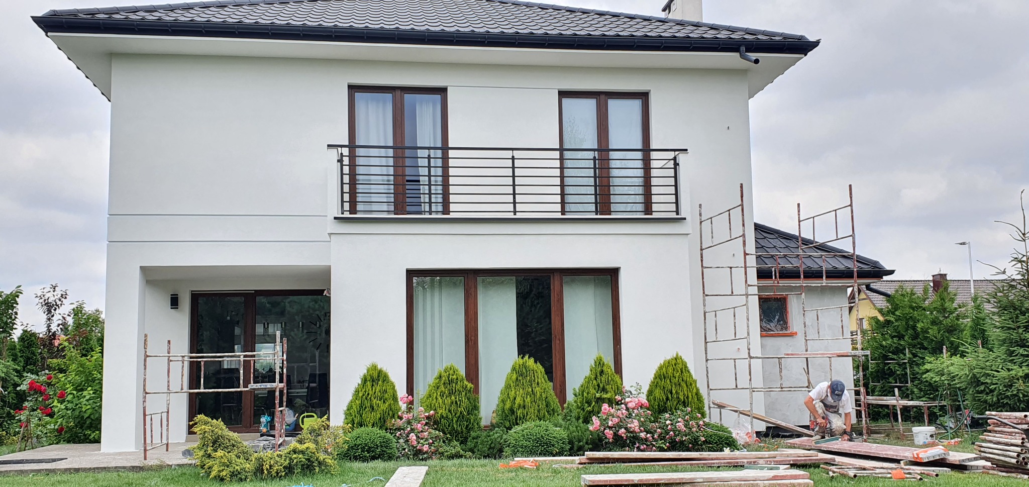 Dwukondygnacyjny dom z białymi ścianami i balkonem z czarną balustradą, widok na elewację z ogrodu, gdzie pracownik przygotowuje elementy rusztowania.
