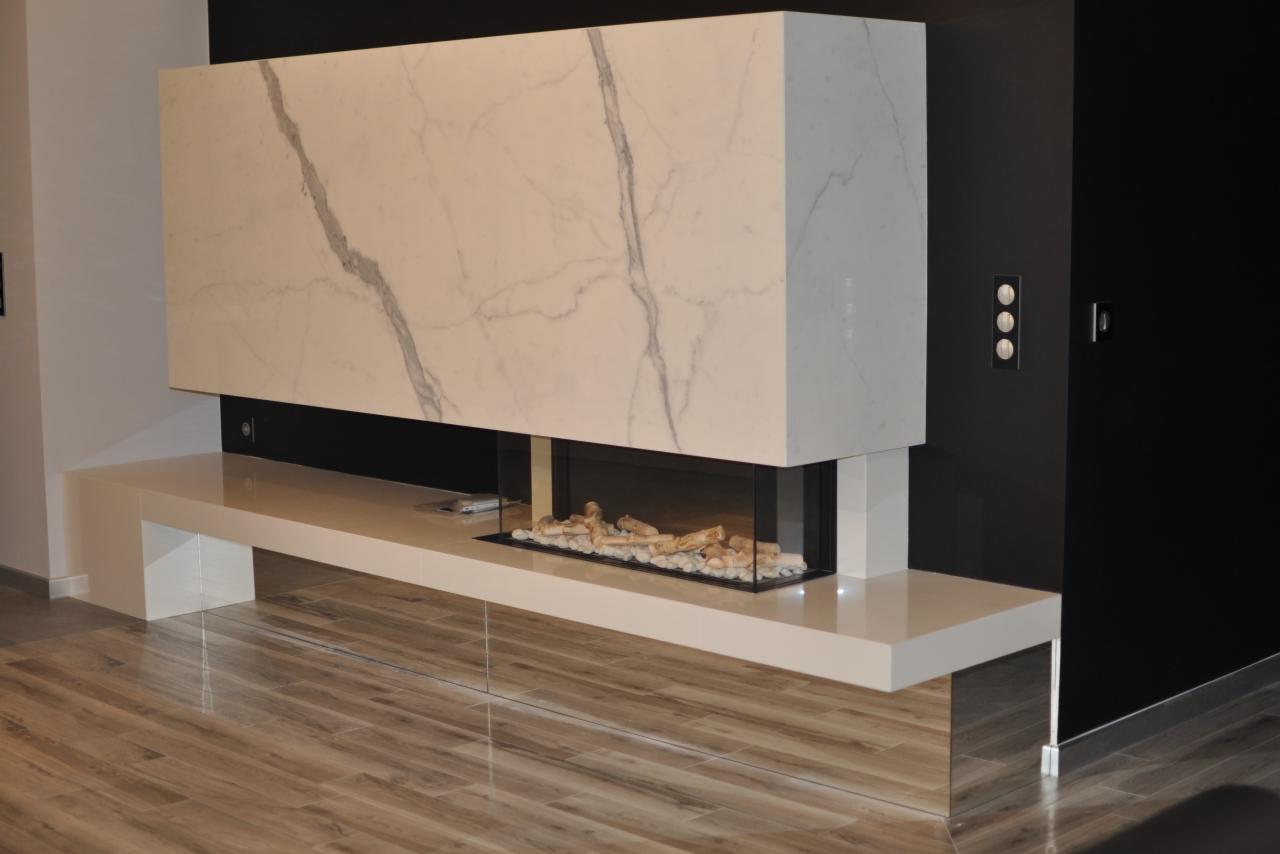 Nowoczesny kominek z obudową z białego marmuru z szarymi żyłami, przeszklony wkład z dekoracyjnymi kamieniami, umieszczony na białym, lakierowanym blacie w minimalistycznym wnętrzu z drewnopodobną...