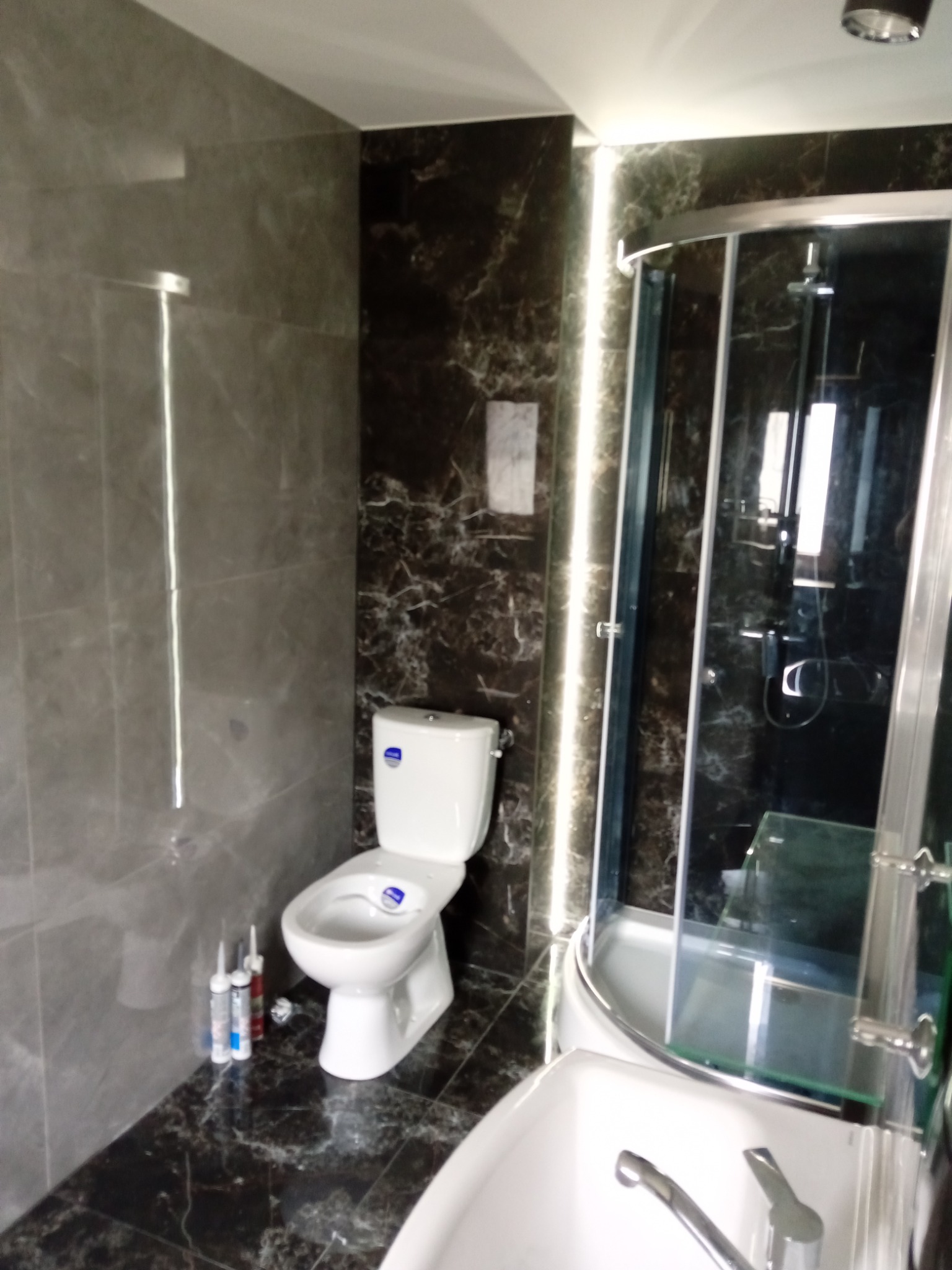 Wykończona łazienka z marmurowymi płytkami, kabiną prysznicową z zaokrąglonym szkłem i białą toaletą. Tubki silikonu leżą na podłodze przy ścianie.