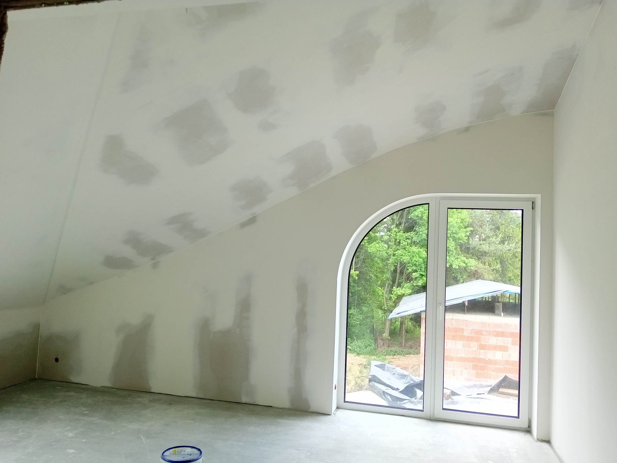 Poddasze w trakcie szpachlowania płyty karton gips, widoczne ślady po szpachli na skośnym suficie, okno z widokiem na budynek w budowie.