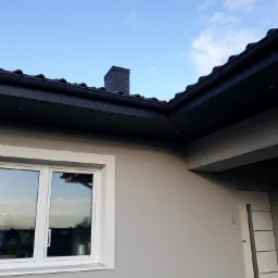 Szary dom jednorodzinny z czarnym dachem i kominem z cegły, widok z dołu na tle błękitnego nieba, białe okno i drzwi wejściowe z szarymi wstawkami.