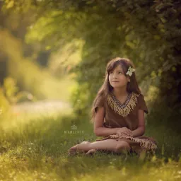 Dziewczynka w brązowej sukience z frędzlami i kwiatem we włosach siedzi na trawie, spoglądając w górę, w tle rozmyte drzewa i światło słoneczne.