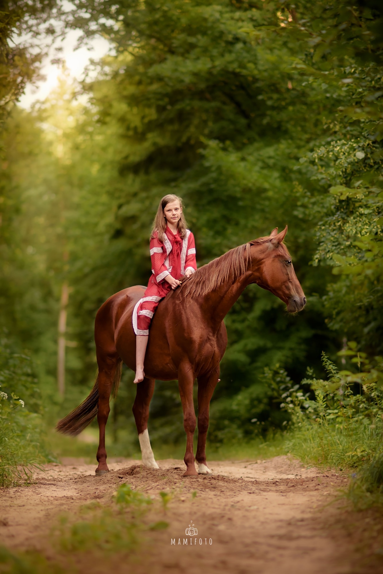 Dziewczynka w czerwonym stroju siedząca okrakiem na brązowym koniu na leśnej ścieżce, z widocznym białym znaczeniem na jednej nodze konia.