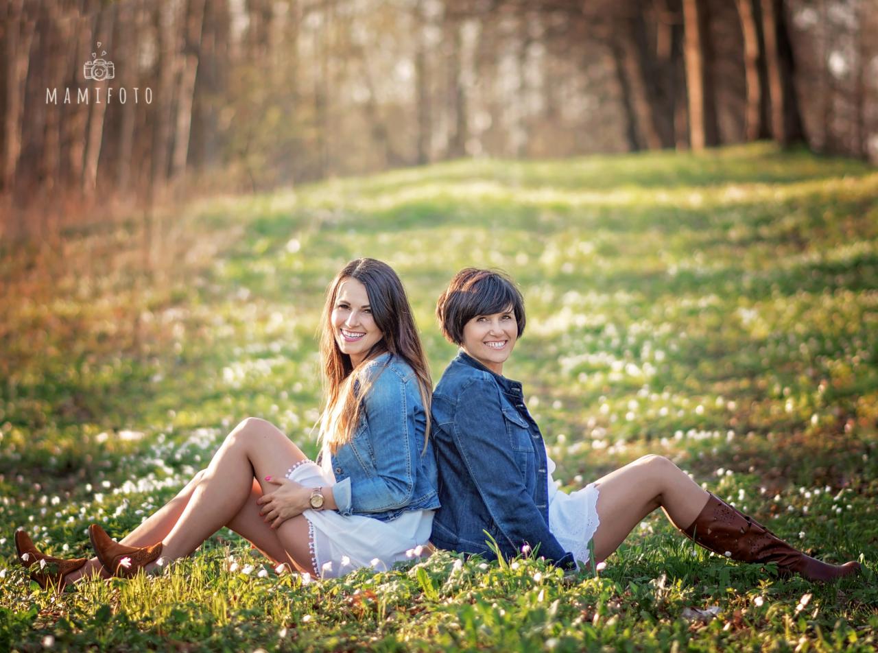 Dwie uśmiechnięte kobiety siedzą plecami do siebie na łące pełnej białych kwiatów, ubrane w jeansowe kurtki i białe sukienki, z widocznymi brązowymi butami kowbojskimi i zegarkiem na ręce jednej...