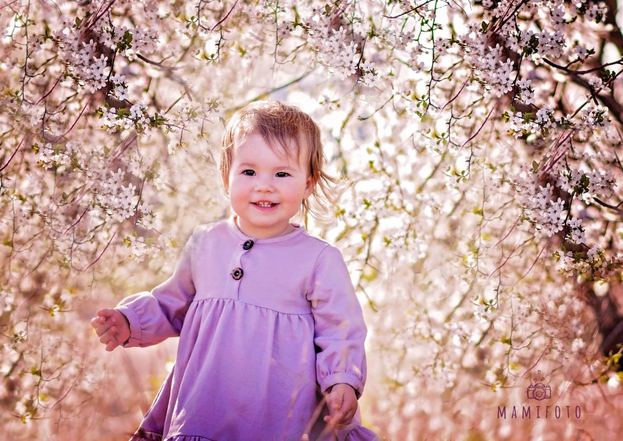 Uśmiechnięta dziewczynka w liliowej sukience stoi na tle kwitnących drzew z białymi kwiatami, z logo fotografa w prawym dolnym rogu.