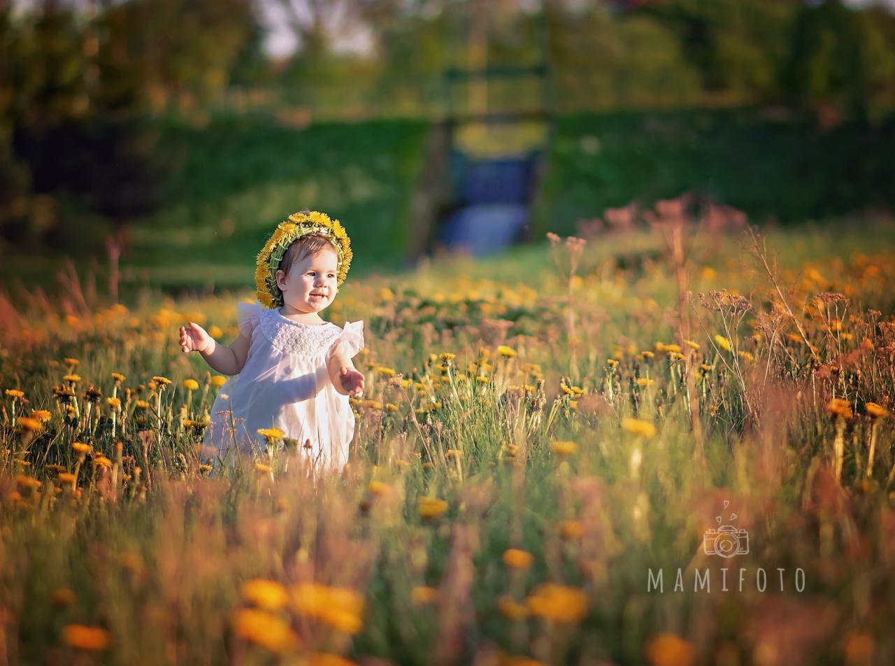 Uśmiechnięta dziewczynka w wianku z żółtych kwiatów, ubrana w białą sukienkę, stoi na łące pełnej dmuchawców, z zamazanym tłem z zielenią i małym mostkiem.