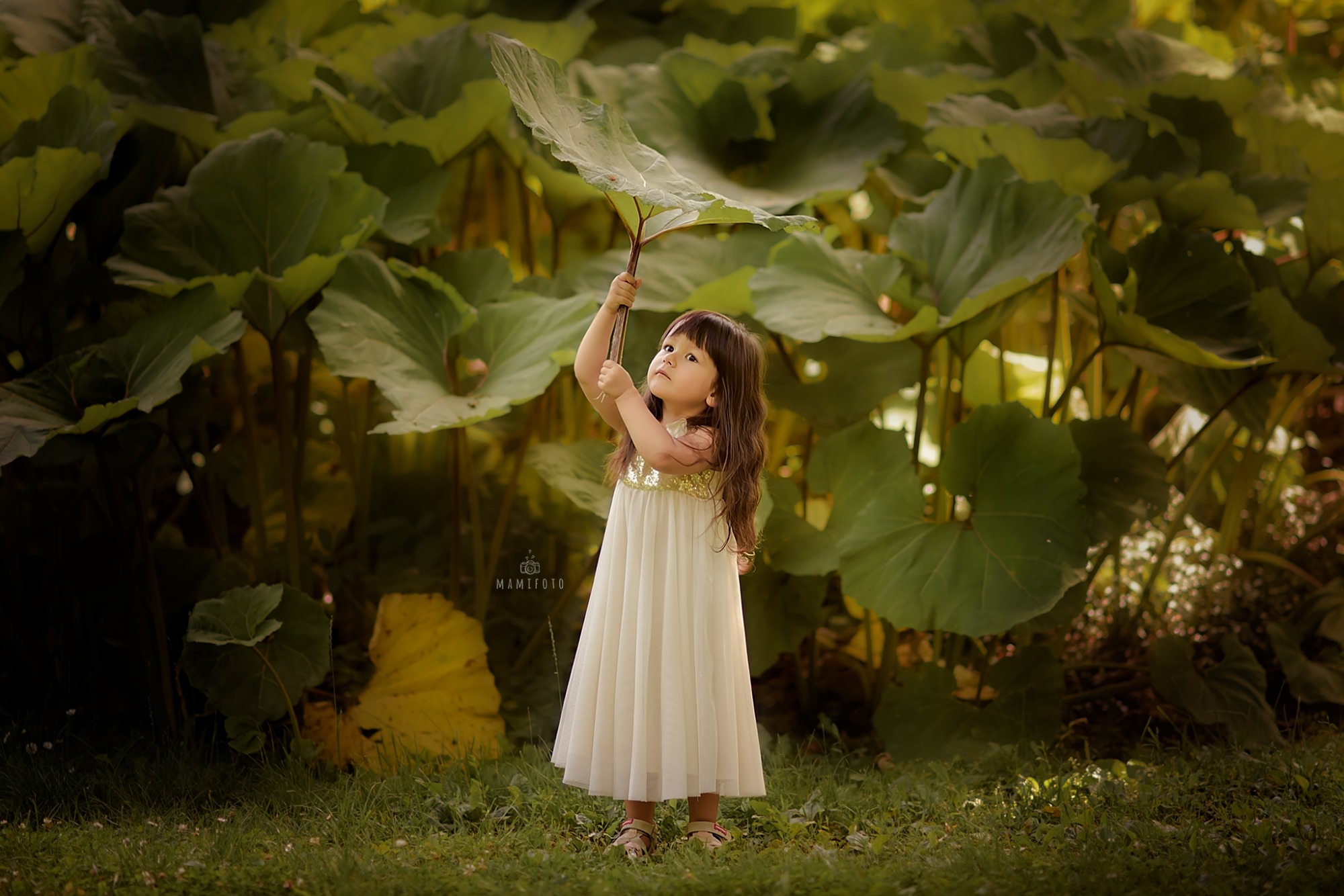 Dziewczynka w białej sukience z cekinami trzyma nad głową duży liść na tle gęstej zieleni liści.