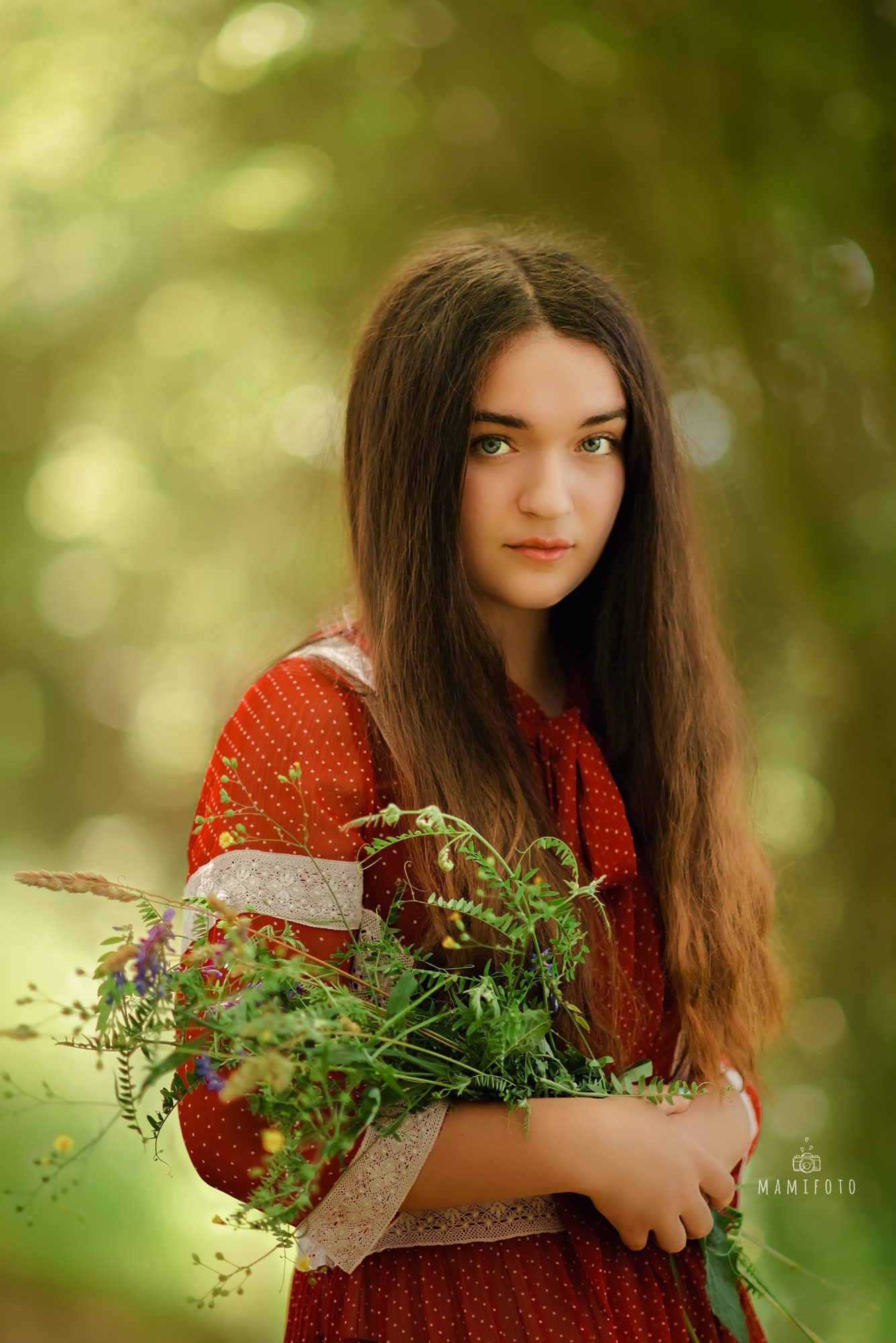 Portret młodej kobiety z długimi brązowymi włosami i zielonymi oczami, trzymającej bukiet polnych kwiatów w czerwonej sukience w białe kropki z koronkowym wykończeniem na tle rozmytej zieleni