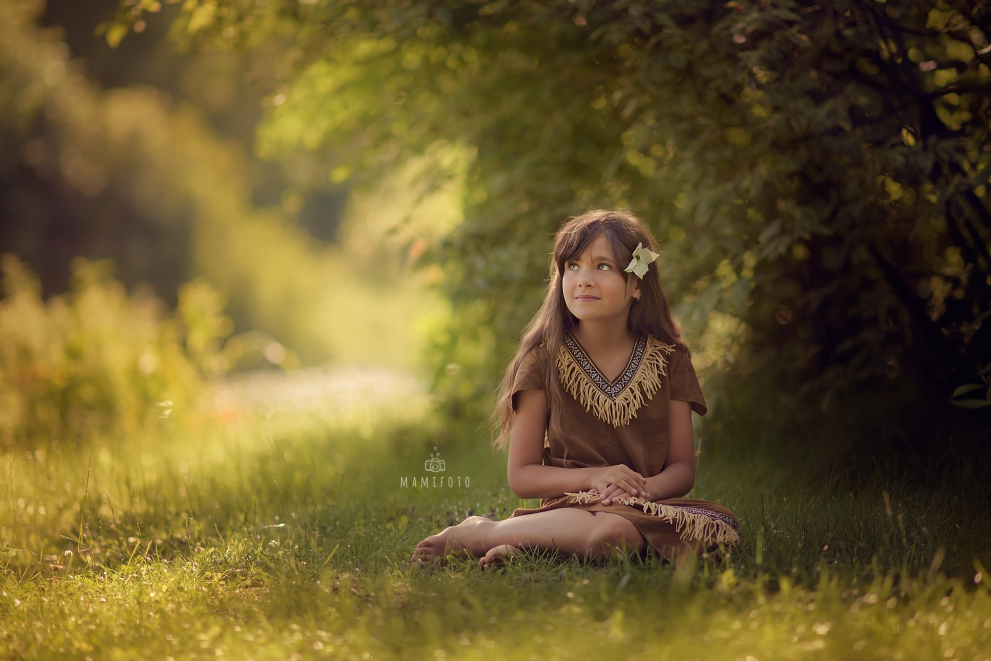 Dziewczynka w brązowej sukience z frędzlami i kwiatem we włosach siedzi na trawie, spoglądając w górę, w tle rozmyte drzewa i światło słoneczne.