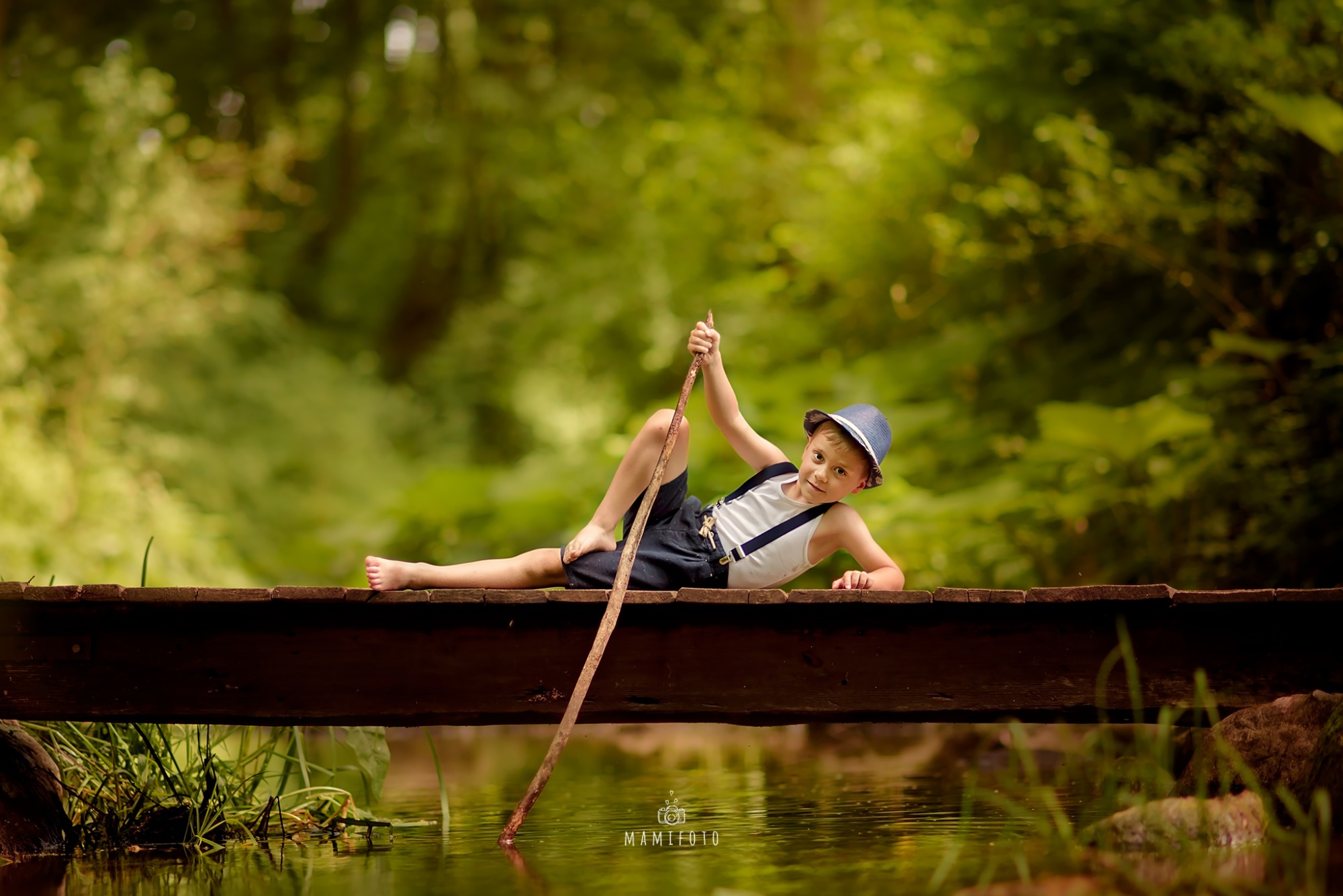 Chłopiec w kapeluszu i szelkach leży na drewnianym mostku nad wodą, trzymając w ręku kij, tło z zieleni