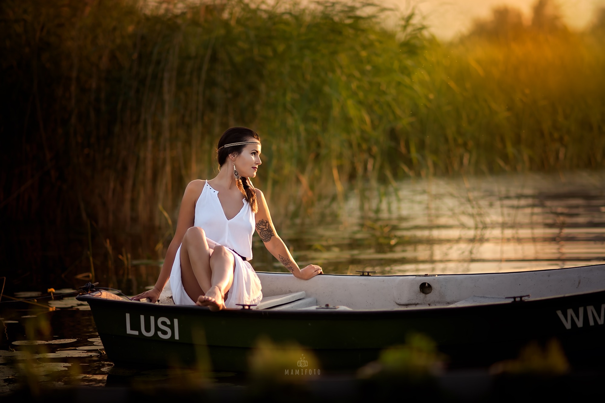 Kobieta w białej sukience siedzi w łódce na tle trzcin i wody o zachodzie słońca, z widocznym tatuażem na ramieniu i opaską na włosach.