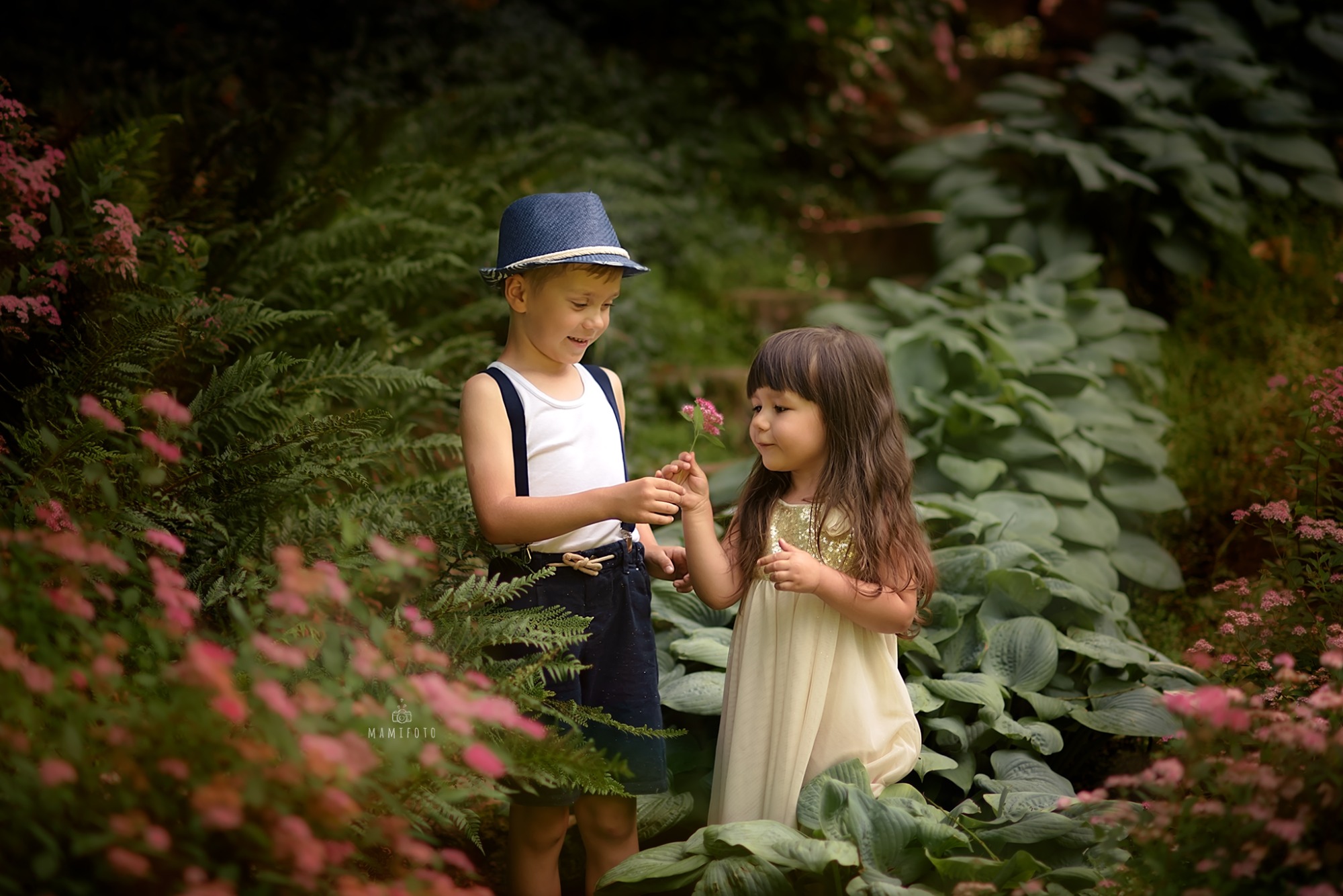 Chłopiec w kapeluszu i dziewczynka w sukience wymieniają się kwiatkiem na tle kwitnącej zieleni w Bartoszycach, uchwycone w miękkim świetle.