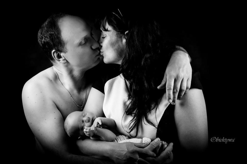 Czarno-białe zdjęcie całującej się pary trzymającej śpiącego noworodka, ciemne tło, delikatne oświetlenie, widoczny łańcuszek na szyi mężczyzny.