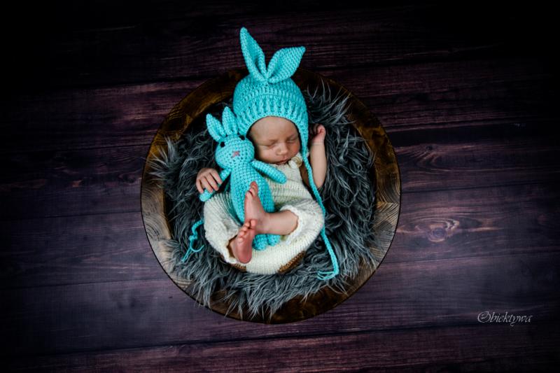 Noworodek śpiący w drewnianej misie wyłożonej szarym futerkiem, ubrany w biały kombinezon i turkusową czapkę z uszami królika, trzymający pluszowego królika w tym samym kolorze.