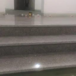 Trzy stopnie schodów z szarego kamienia, z widocznymi elementami poziomowania na podłodze w tle, sugerujące trwający montaż.