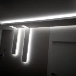 Nowoczesne oświetlenie sufitowe z pionowymi panelami LED na ścianie w pomieszczeniu w Nowym Targu, widoczna biała ściana i drzwi.