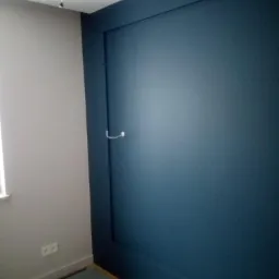 Ściana pomalowana na granatowo z dekoracyjną ramą, widoczny fragment szarej ściany z gniazdkiem elektrycznym i surowa podłoga z plamami farby.