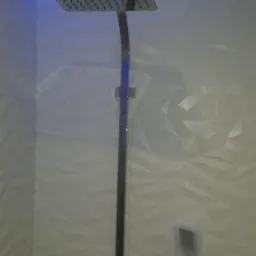 Kwadratowa deszczownica chromowana zamontowana na ścianie z trójwymiarowymi białymi płytkami, podświetlenie LED w suficie.