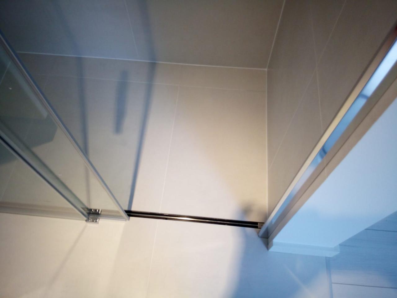 Szczegółowe ujęcie kabiny prysznicowej z przesuwnymi szklanymi drzwiami, zamontowanej w rogu wyłożonym jasnymi, kwadratowymi płytkami ceramicznymi, widoczne metalowe elementy mocujące i prowadnice.