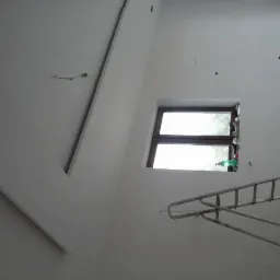 Świeżo pomalowane białe wnętrze z nietypowym sufitem, drabina oparta o ścianę pod oknem, widoczne ślady po remoncie na podłodze, puszka farby w rogu.