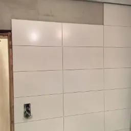 Ściana wyłożona białymi, matowymi płytkami ceramicznymi w trakcie montażu, widoczne kable wystające z puszki elektrycznej, surowy sufit w tle.