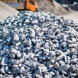 KRISPOL Krzysztof Niedziałek - Kupa jasnoszarego żwiru na placu budowy, z rozmytym pomarańczowym pojazdem w tle. Drobny żwir o nieregularnych kształtach, oświetlony naturalnym światłem.