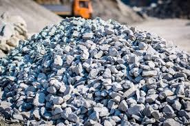 Kupa jasnoszarego żwiru na placu budowy, z rozmytym pomarańczowym pojazdem w tle. Drobny żwir o nieregularnych kształtach, oświetlony naturalnym światłem.