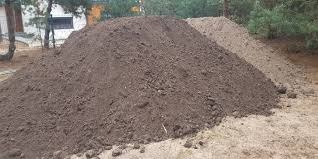 Dwie pryzmy ziemi ogrodowej o różnej barwie, ciemniejsza z przodu i jaśniejsza w tle, na piaszczystym podłożu, w otoczeniu drzew i budynku.