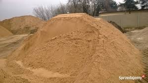 Kupa piasku budowlanego o jasnobrązowej barwie, uformowana w stożek, na tle szarego nieba i fragmentu betonowego muru. Widoczne ziarna piasku.