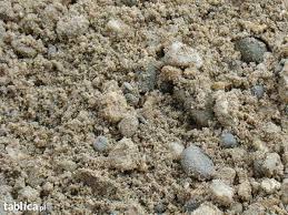 Piasek budowlany z widocznymi kamyczkami o różnej granulacji, oświetlony naturalnym światłem, wypełniający kadr. Tekstura piasku wyraźnie widoczna.