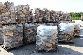 Stosy worków z pociętym drewnem ułożonych na paletach, część worków przezroczysta, część owinięta folią, widoczne jasne i ciemne kawałki drewna, w tle zieleń drzew.