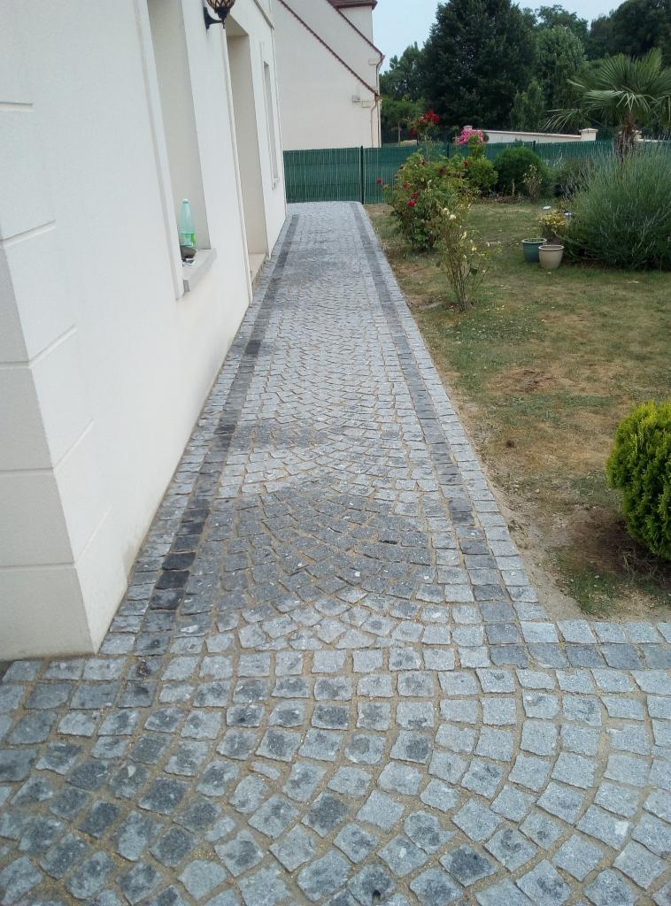 Ścieżka z szarej kostki granitowej ułożonej w wachlarzowy wzór, obrzeża z ciemniejszej kostki, prowadząca wzdłuż białego domu z widocznym ogrodem w tle.