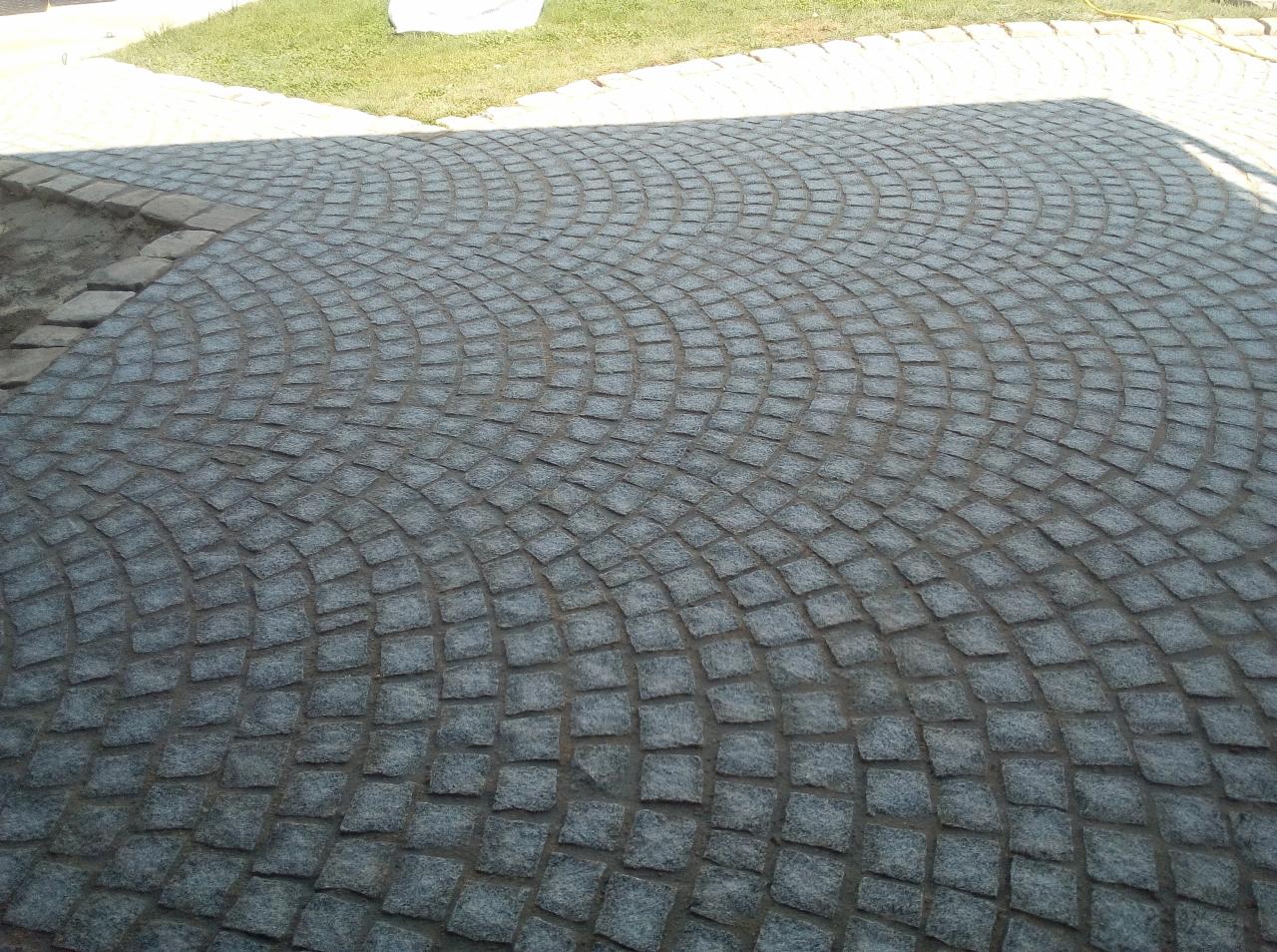 Szara kostka granitowa ułożona w łukowaty wzór na podjeździe, z fragmentem trawnika w tle.