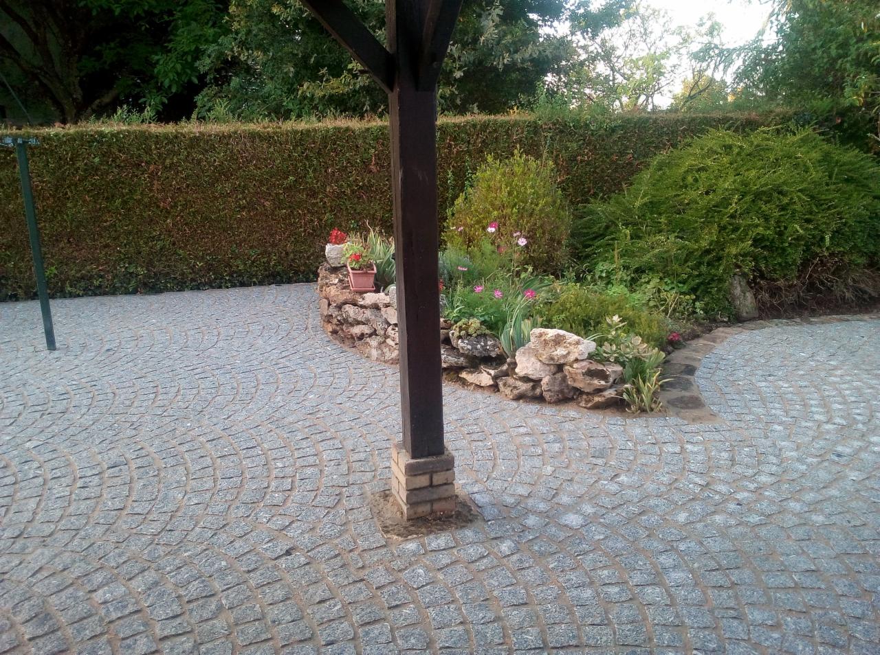 Szara kostka granitowa ułożona w łukowaty wzór, obok rabata z kwiatami i krzewami, widoczny element drewnianej konstrukcji z ceglaną podstawą.