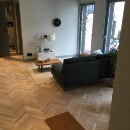 Slazyk BYGG - Salon z parkietem ułożonym we wzór jodełki, szarą sofą i widokiem na zewnątrz przez duże okna.